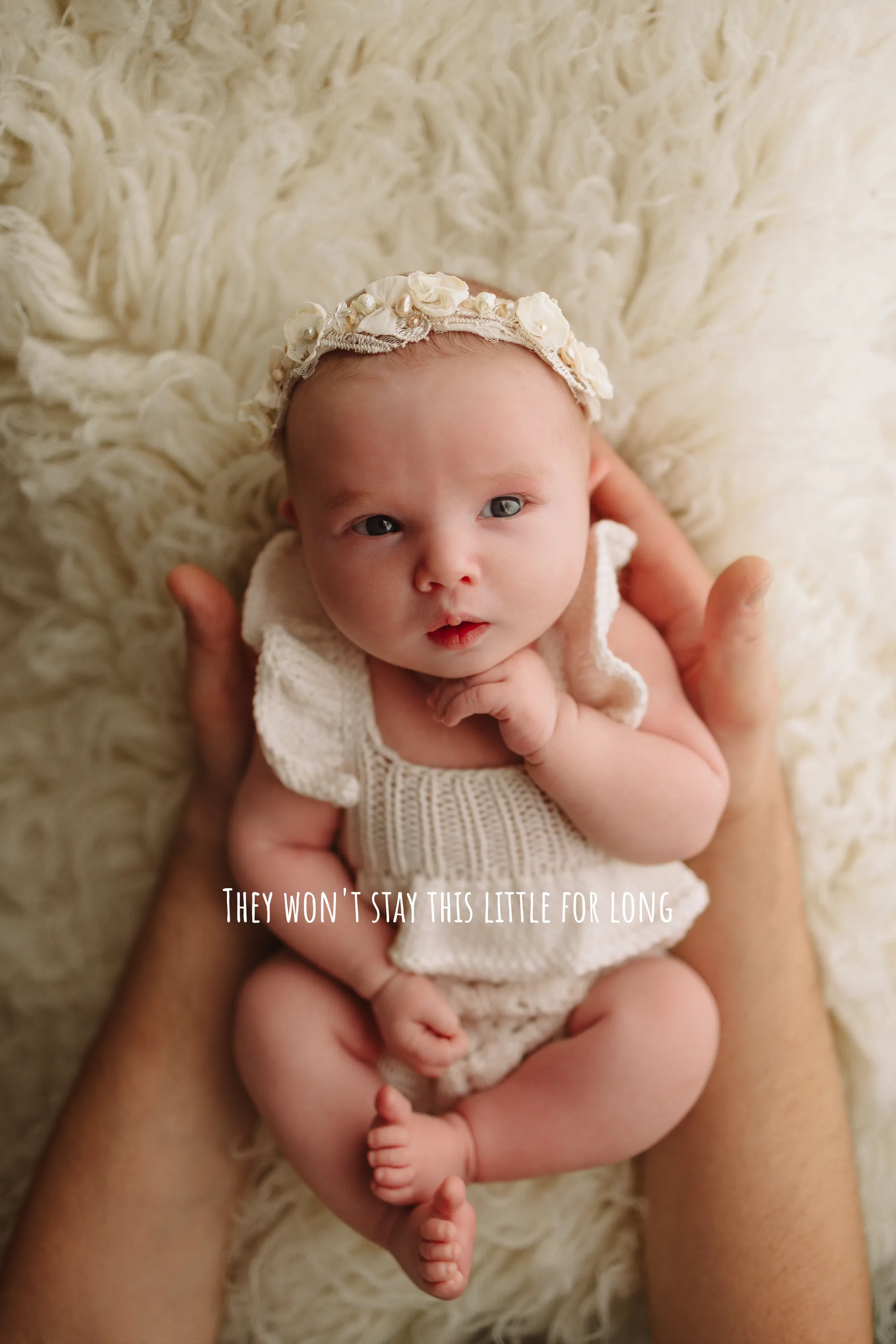 Newborn baby girl in a cream knit romper and floral headband, cradled in parent's hands on sheepskin at our West Wickham studio