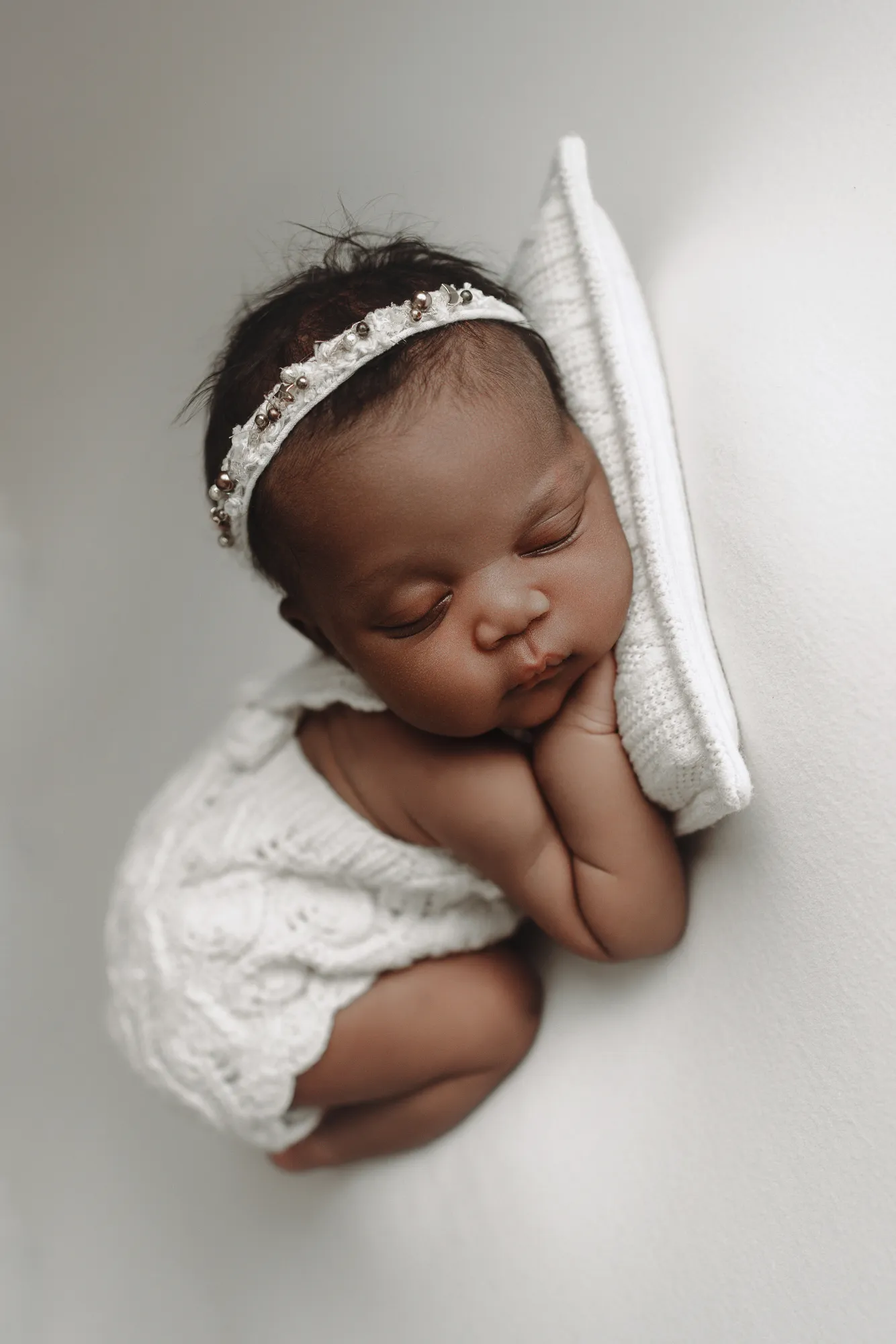 Newborn girl in a white knit romper and pearl headband asleep on a white pillow
