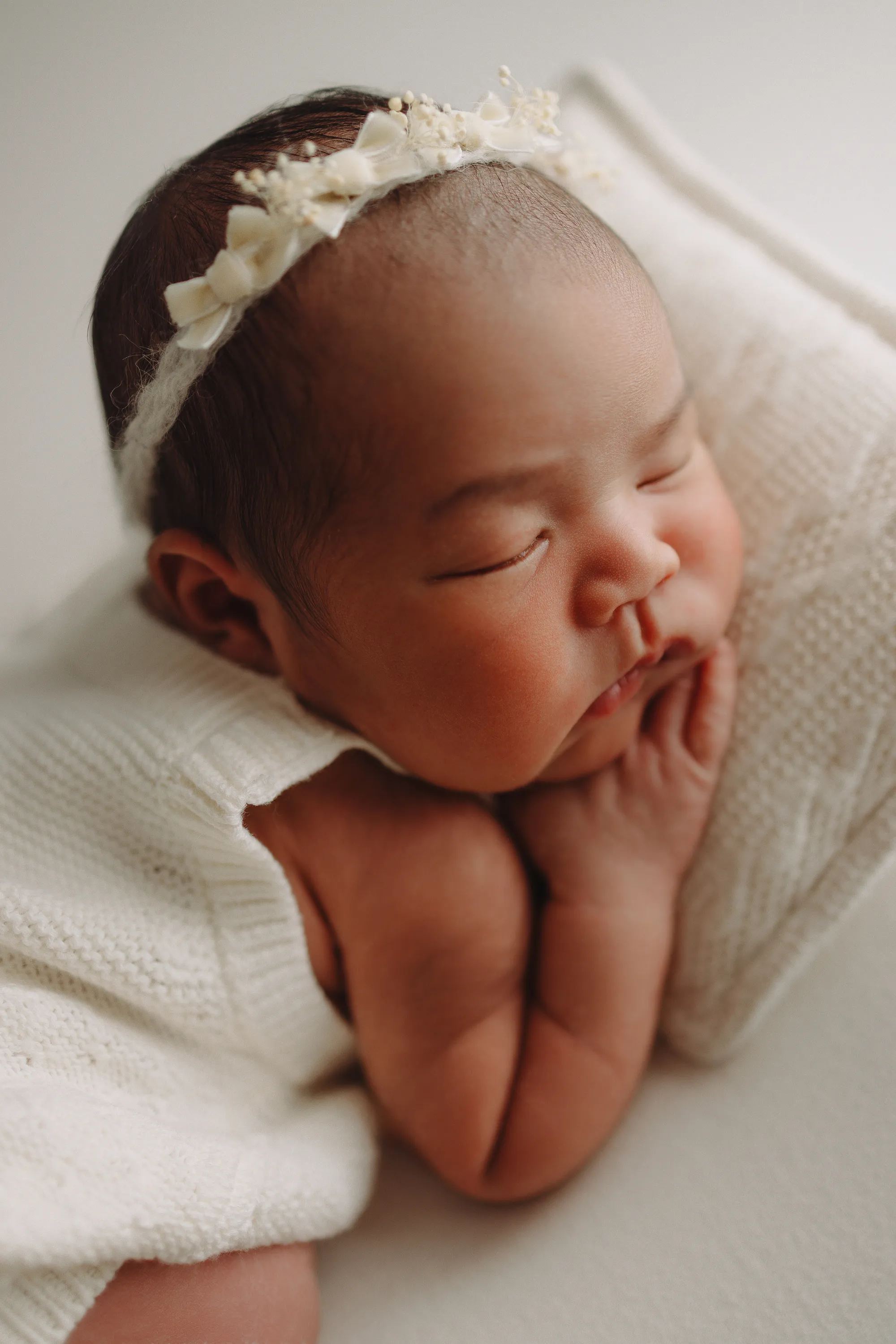 Close-up of a sleeping newborn resting her chin on her hands, wearing a lace headband