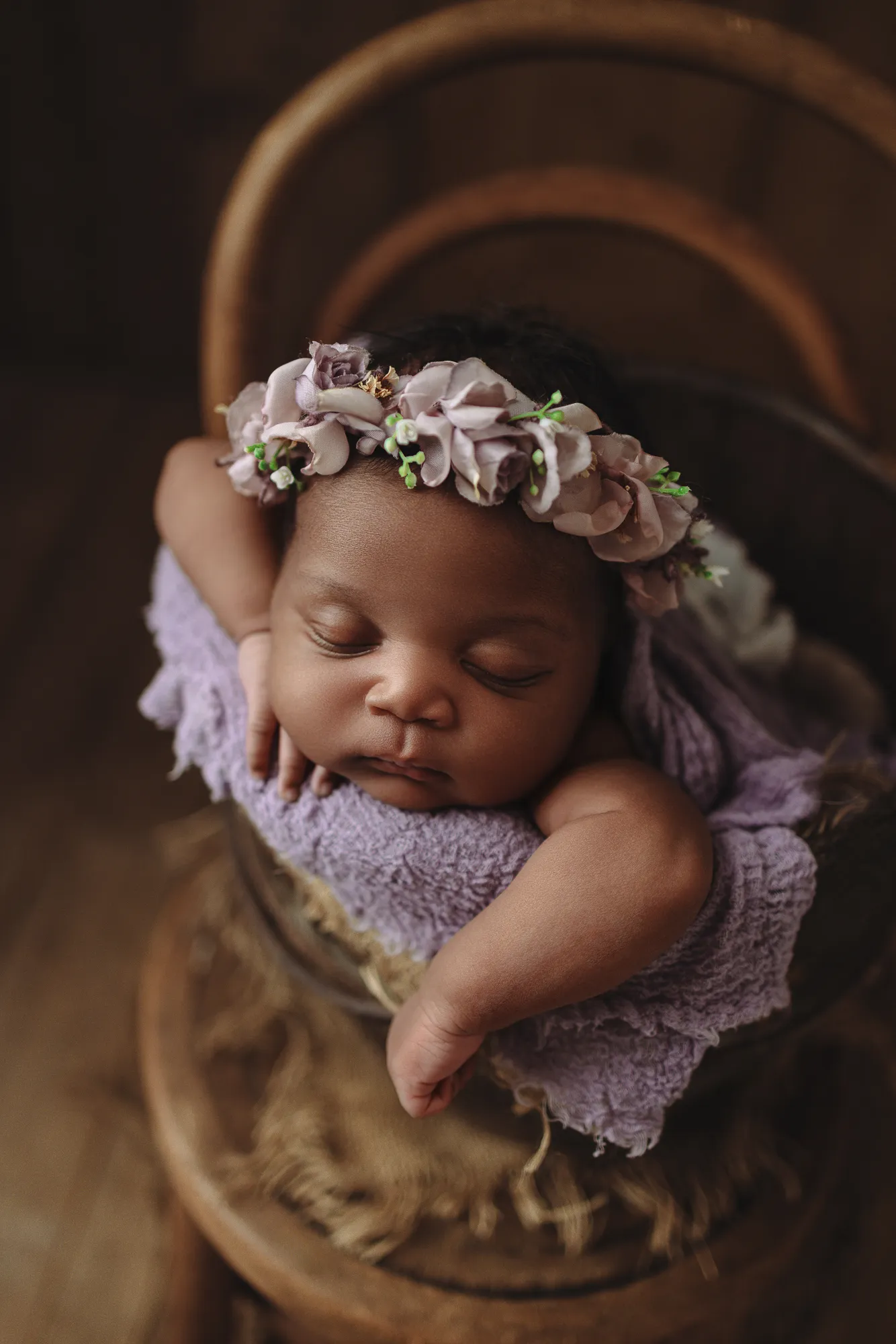 Newborn with a lilac flower crown resting her chin on her arms in a wooden bowl