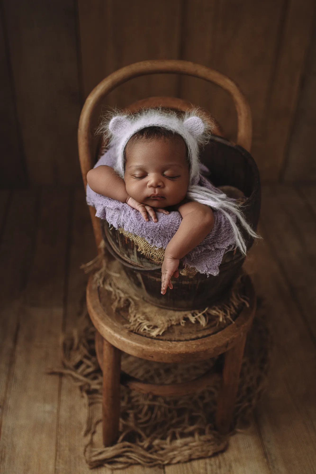 Newborn in a teddy-ear bonnet sleeping in a basket on a vintage wooden chair