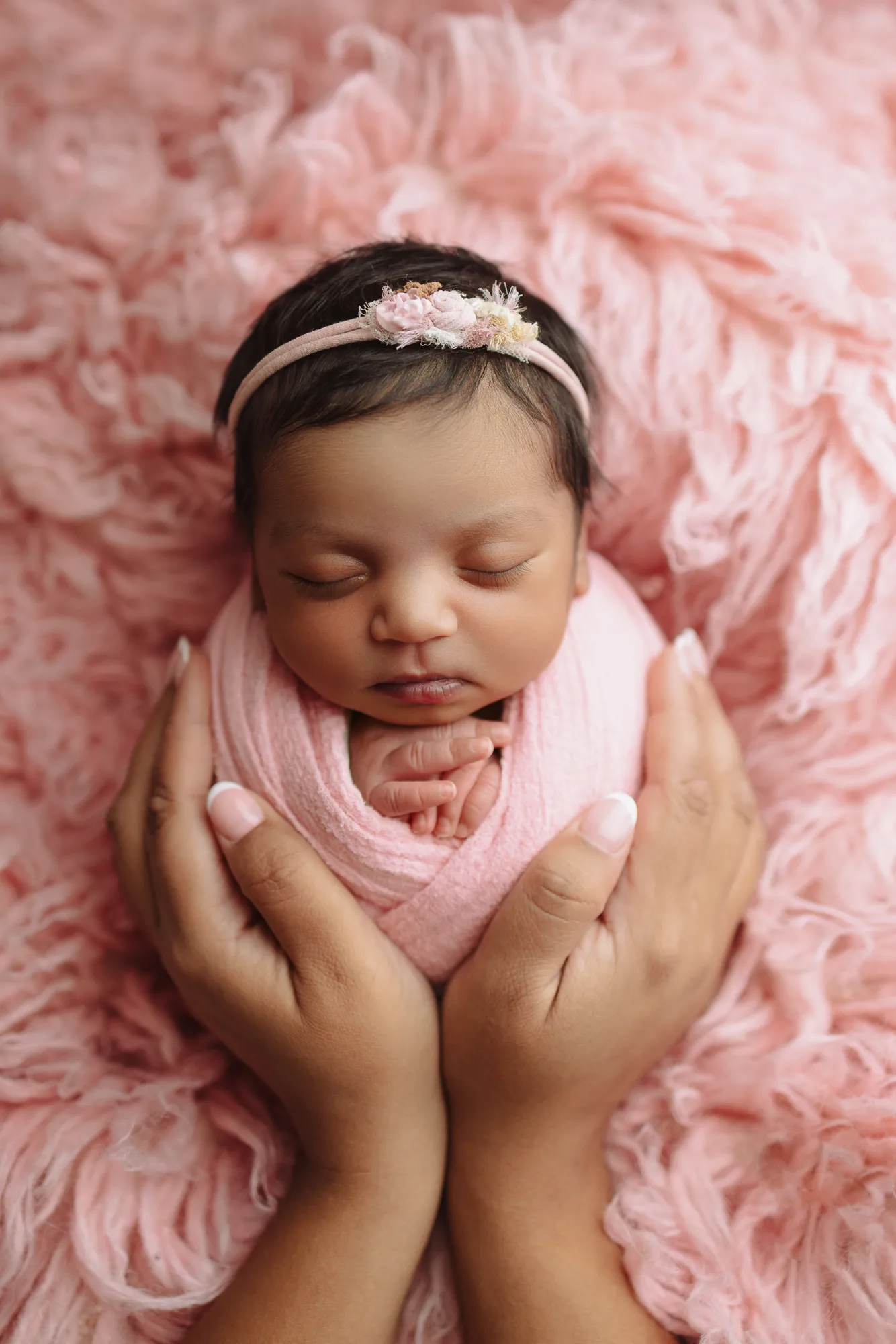 Newborn swaddled in pink, cupped in a parent's hands on pink fur