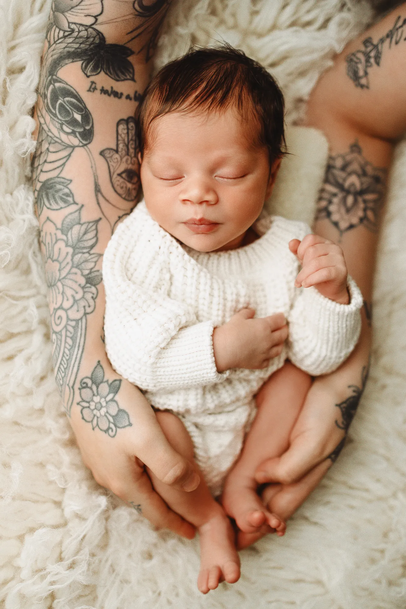 Newborn in a white knit jumper held by a parent with tattooed arms
