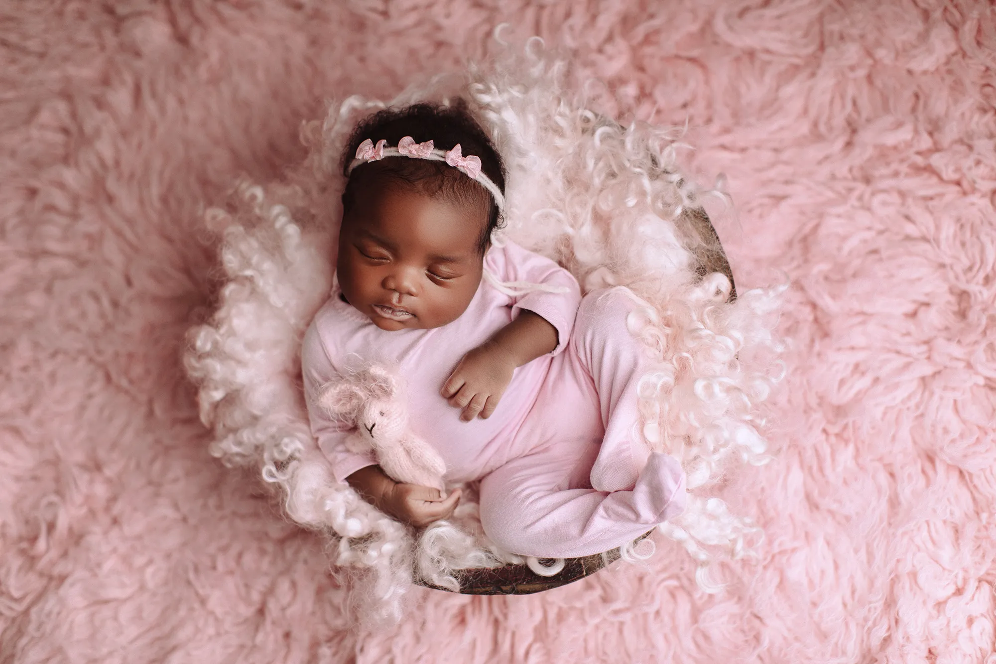 Newborn in a pink romper and headband lying in a bowl on pink fur