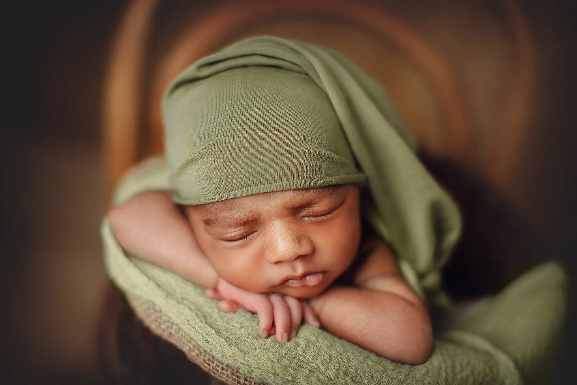 Close-up of a newborn in a sage green sleepy hat resting on folded arms