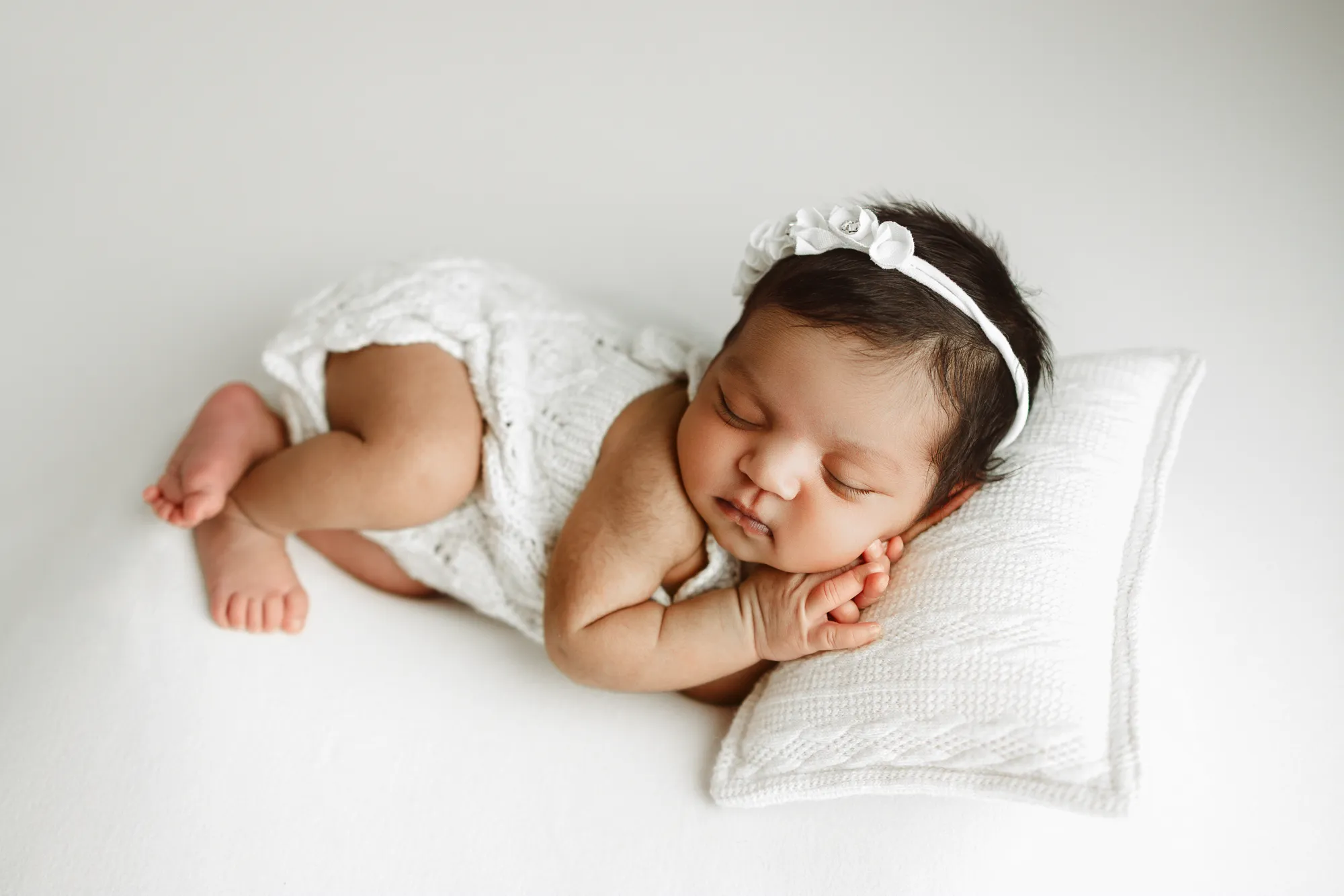 Newborn in a white lace romper sleeping sideways on a white pillow