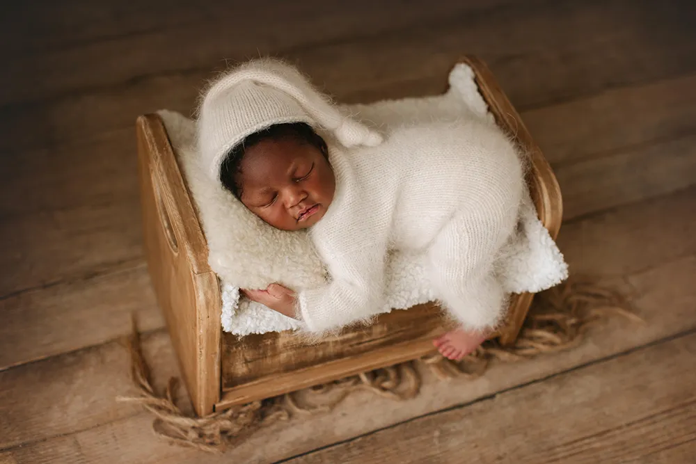 Newborn in a white knit bonnet and romper asleep in a wooden bed