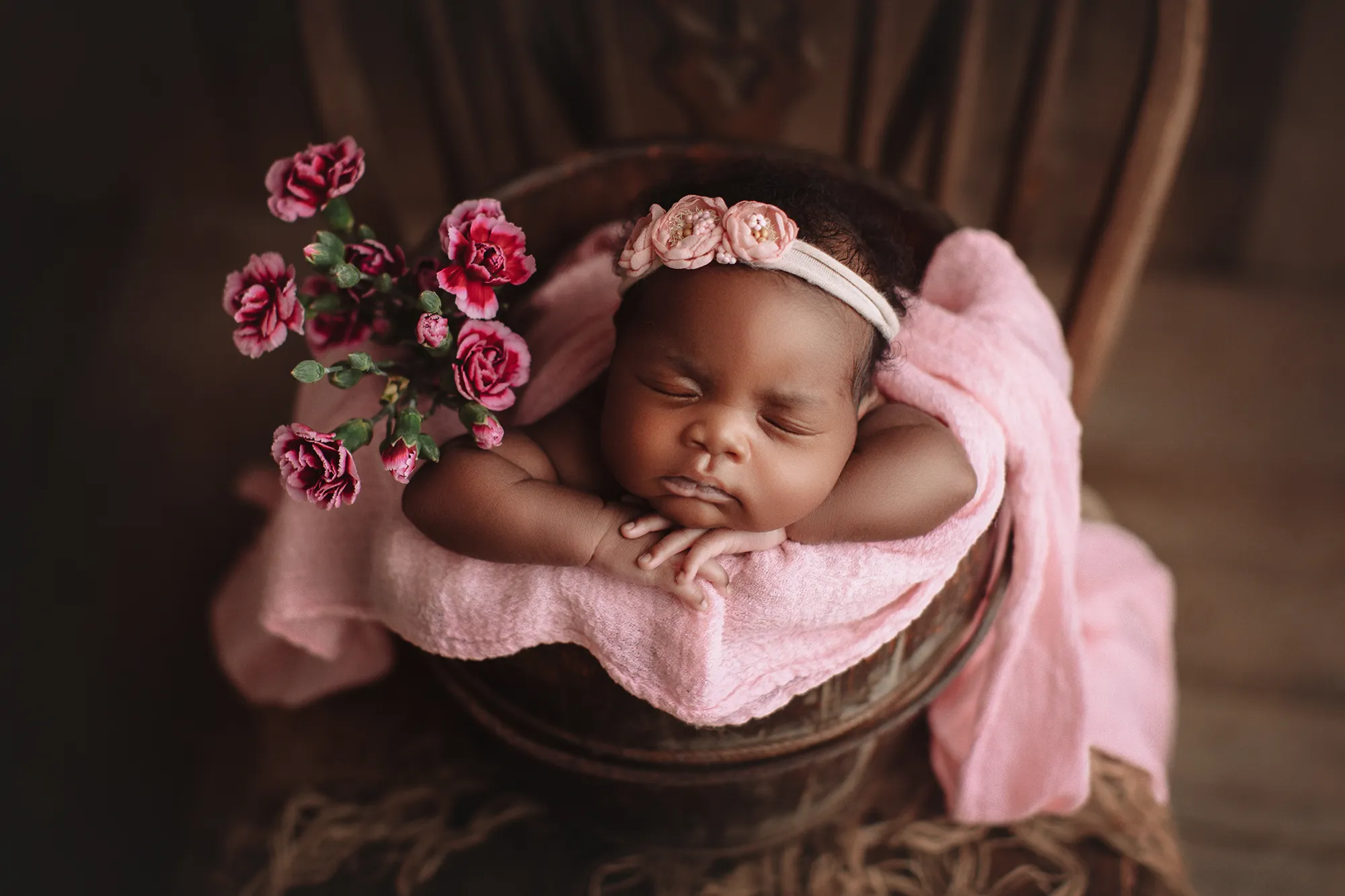 Newborn in a pink wrap asleep in a bucket beside a bunch of pink roses