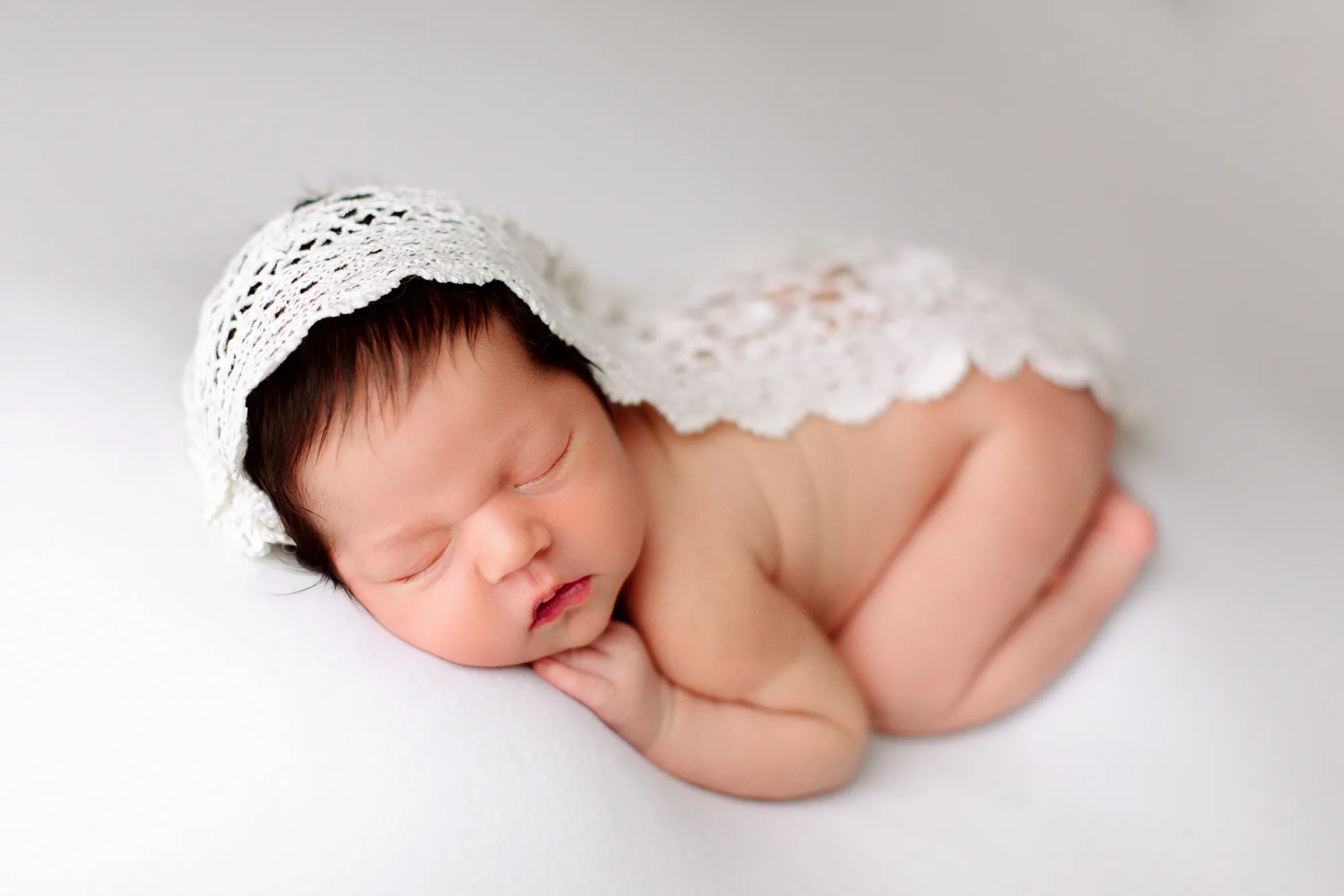 Newborn in a lace bonnet curled up asleep on a white backdrop