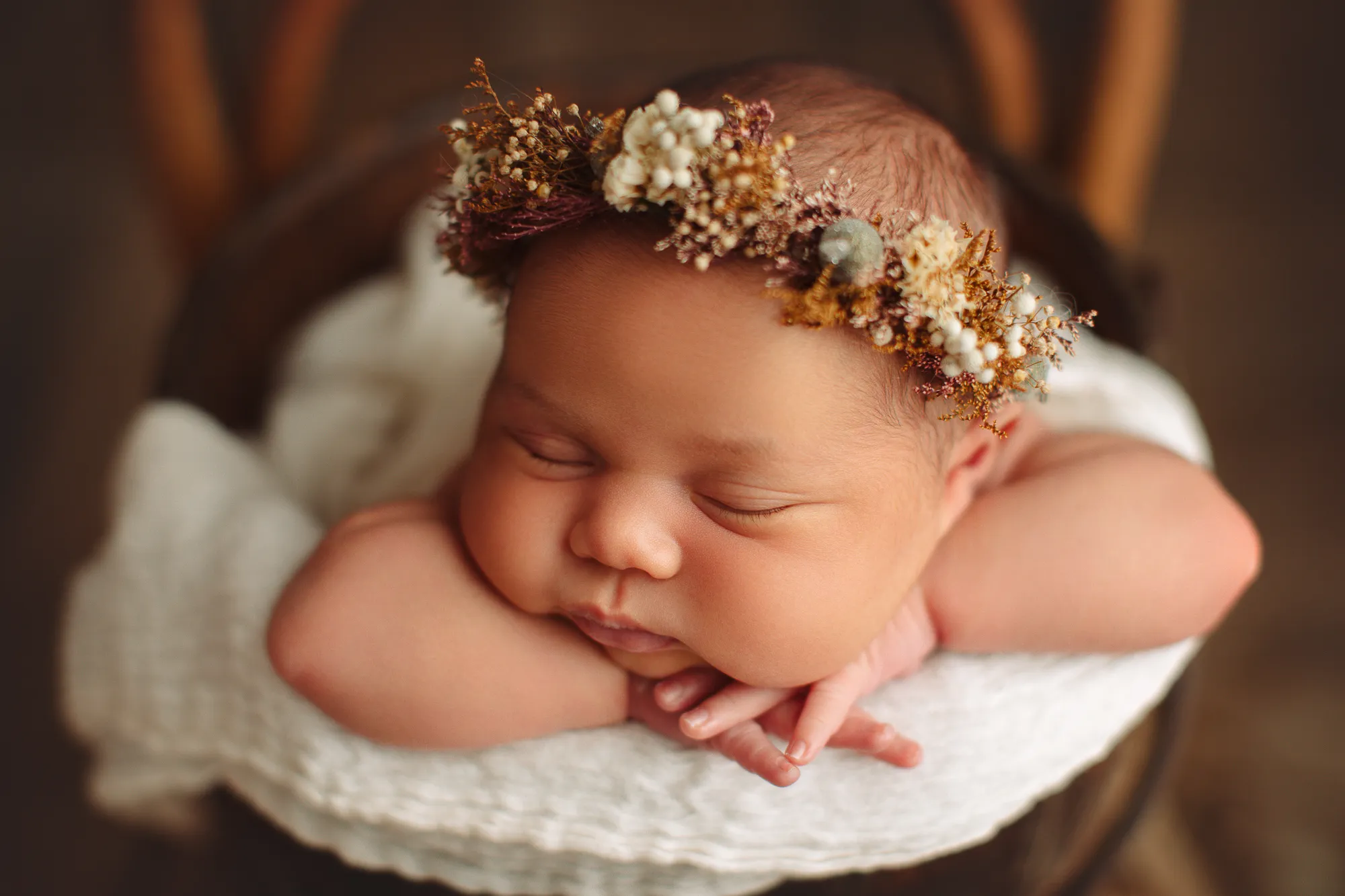 Newborn in a dried-flower crown resting chin on hands in a white-lined bowl