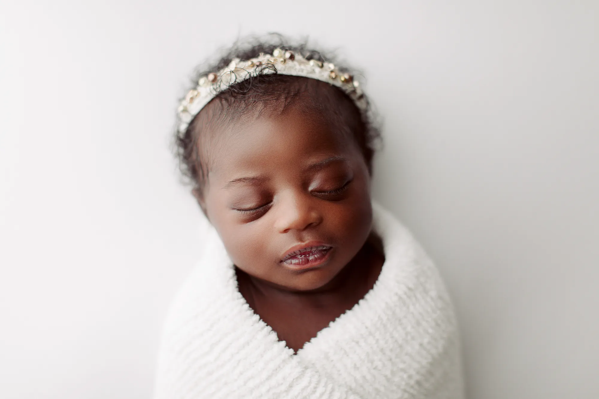 Newborn wrapped in white with a jewelled headband against a white backdrop