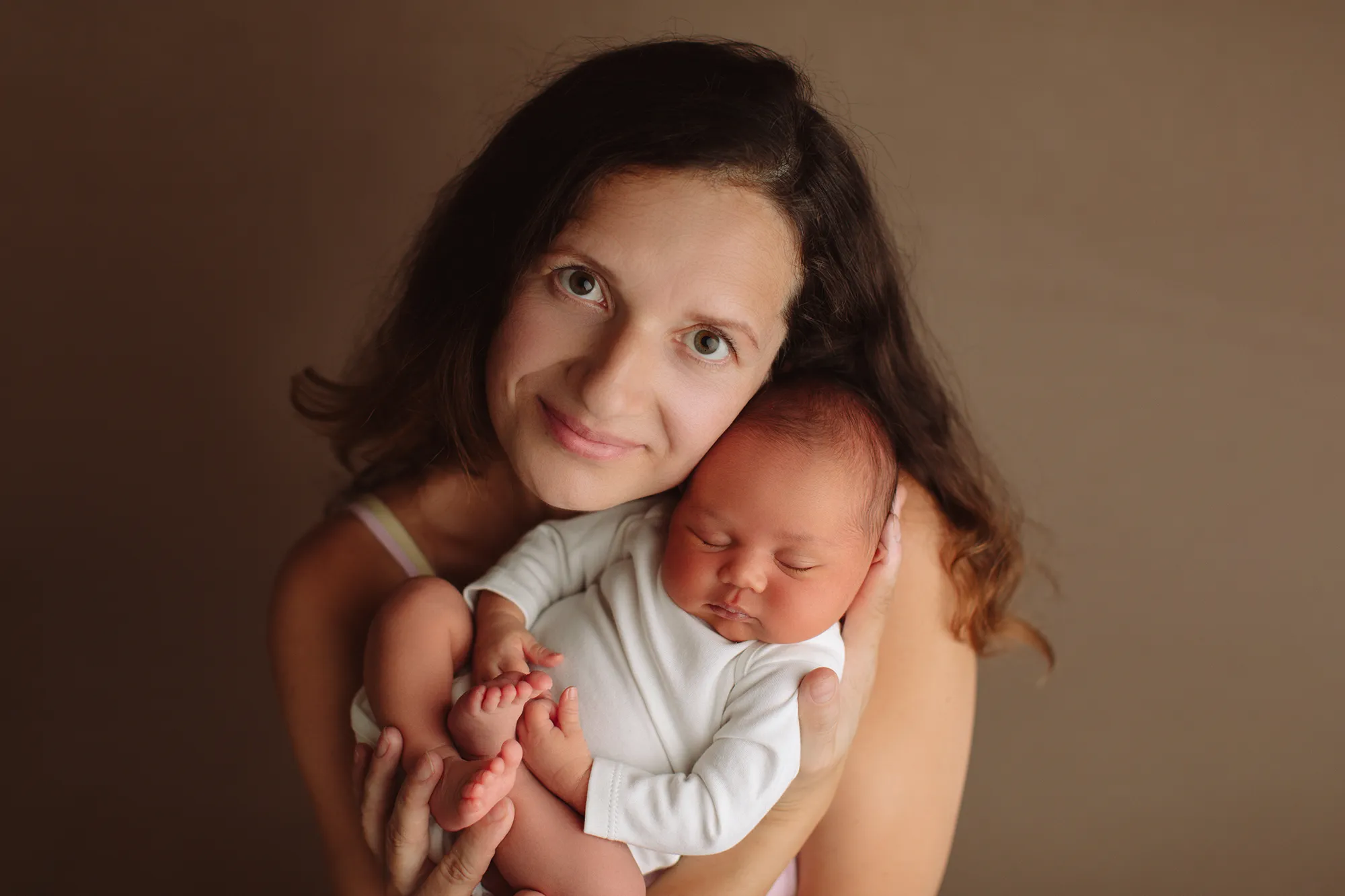 Smiling mother holding her newborn in a white romper against a brown backdrop