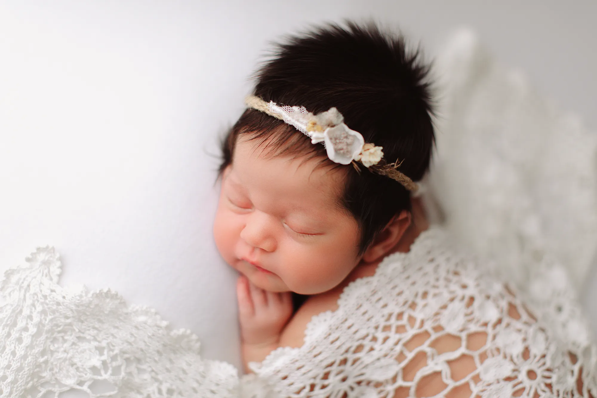 Newborn girl with a floral headband asleep under a white crochet blanket