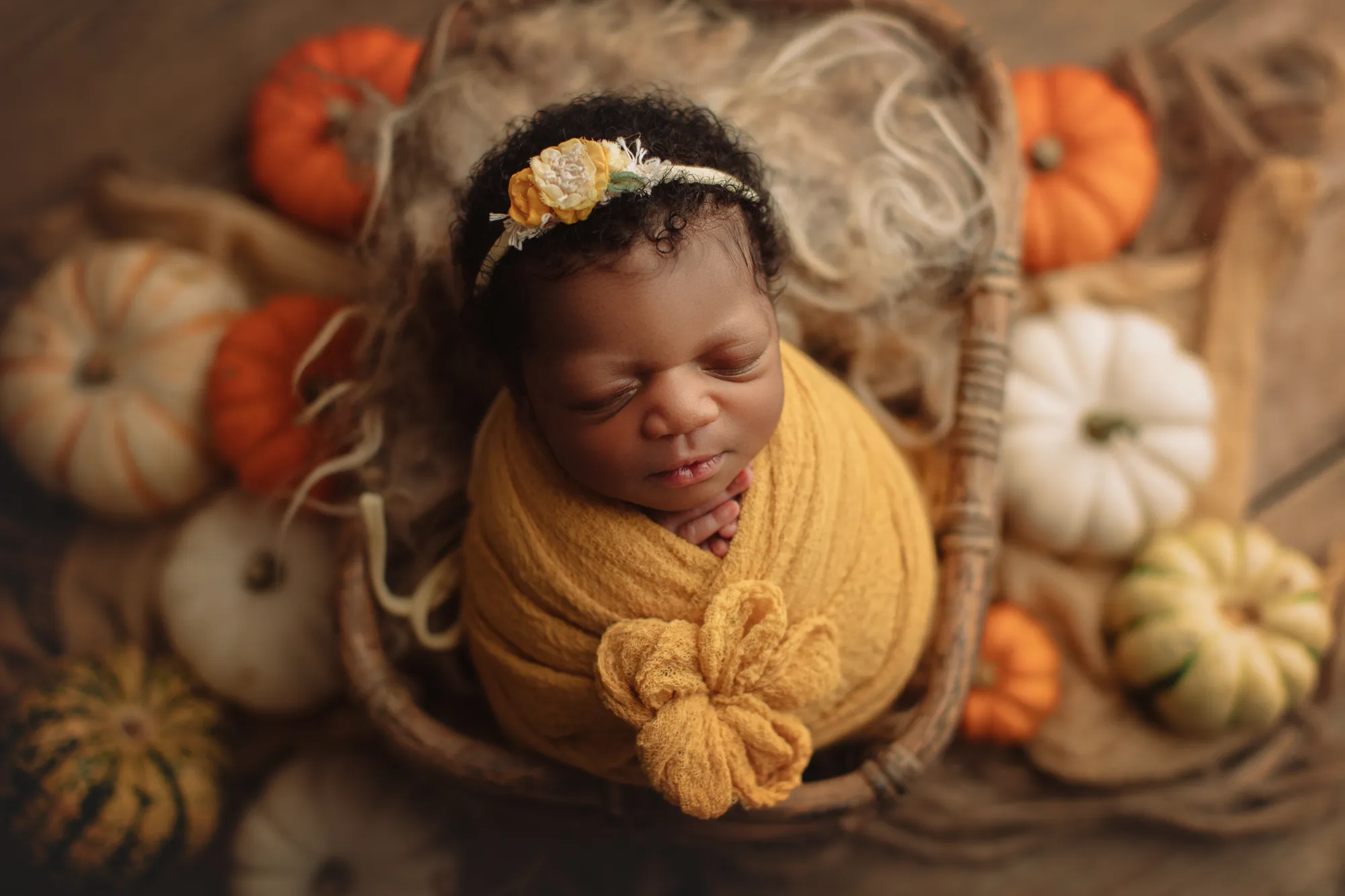 Newborn in a mustard wrap asleep in a basket surrounded by autumn pumpkins