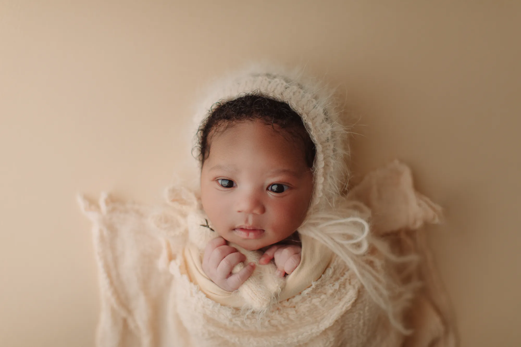 Wide-eyed newborn in a fluffy cream bonnet wrapped in cream knit