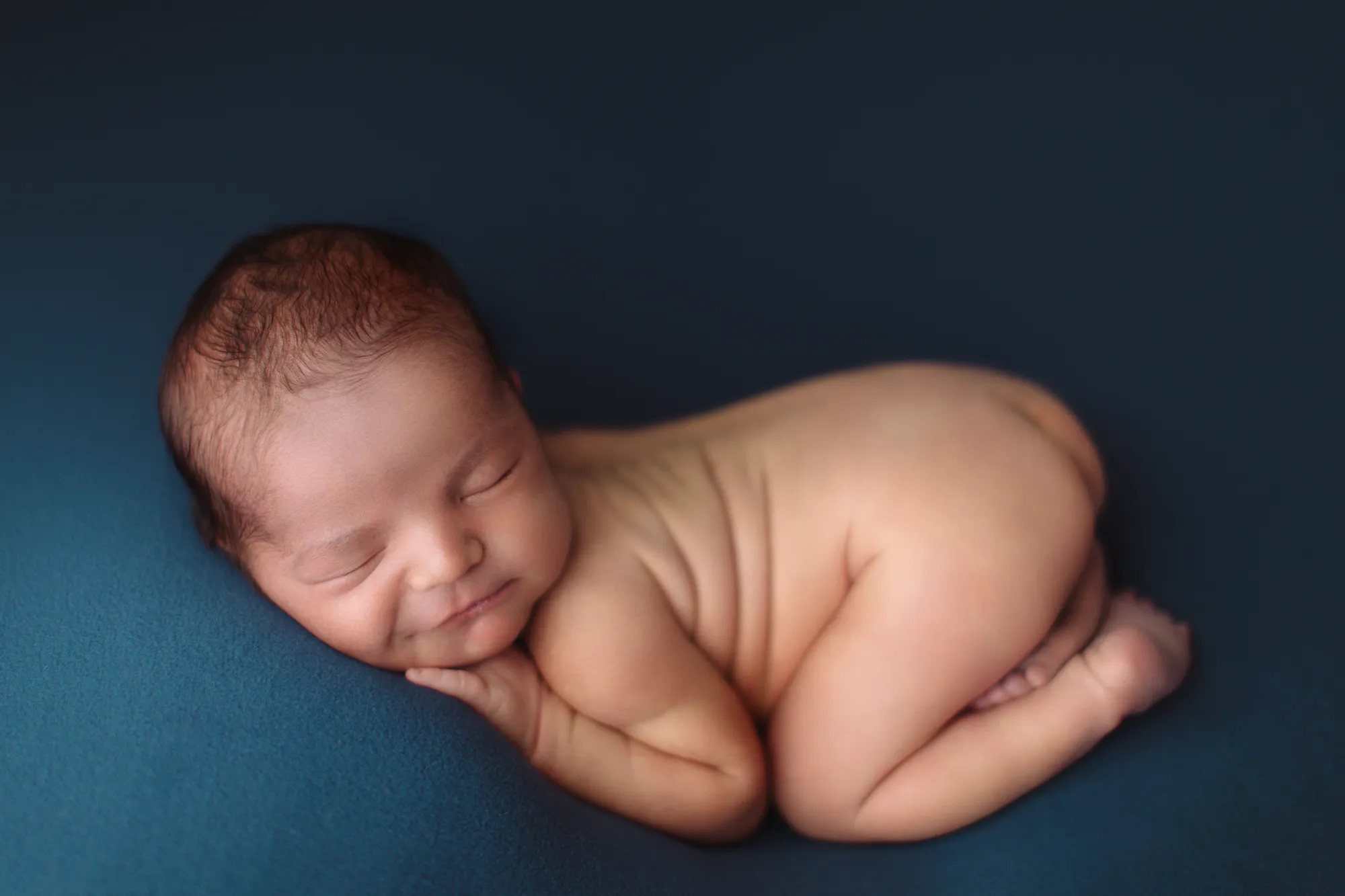 Newborn curled up asleep on a deep blue backdrop