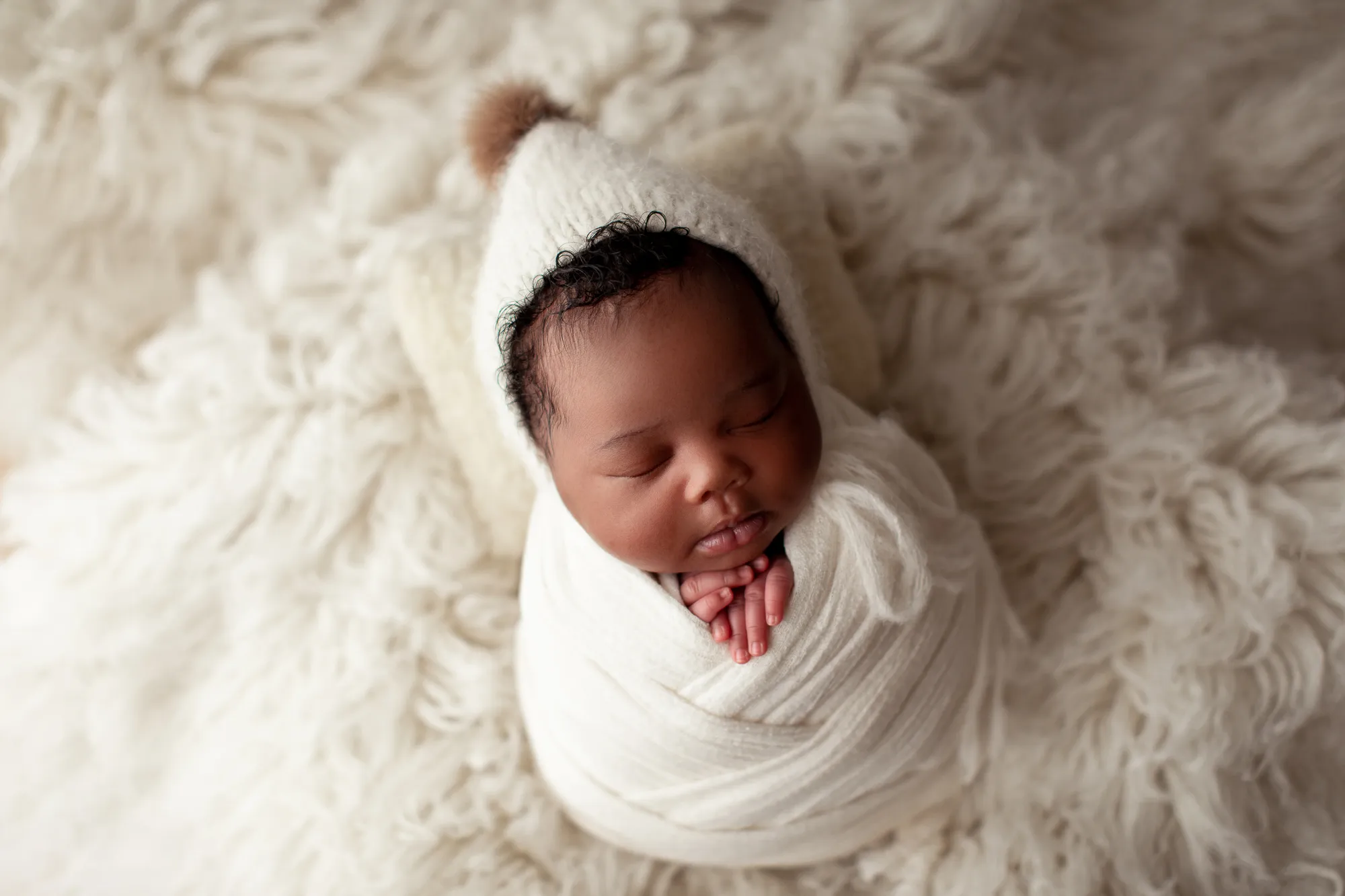 Newborn in a cream knit bonnet swaddled in white on cream fur