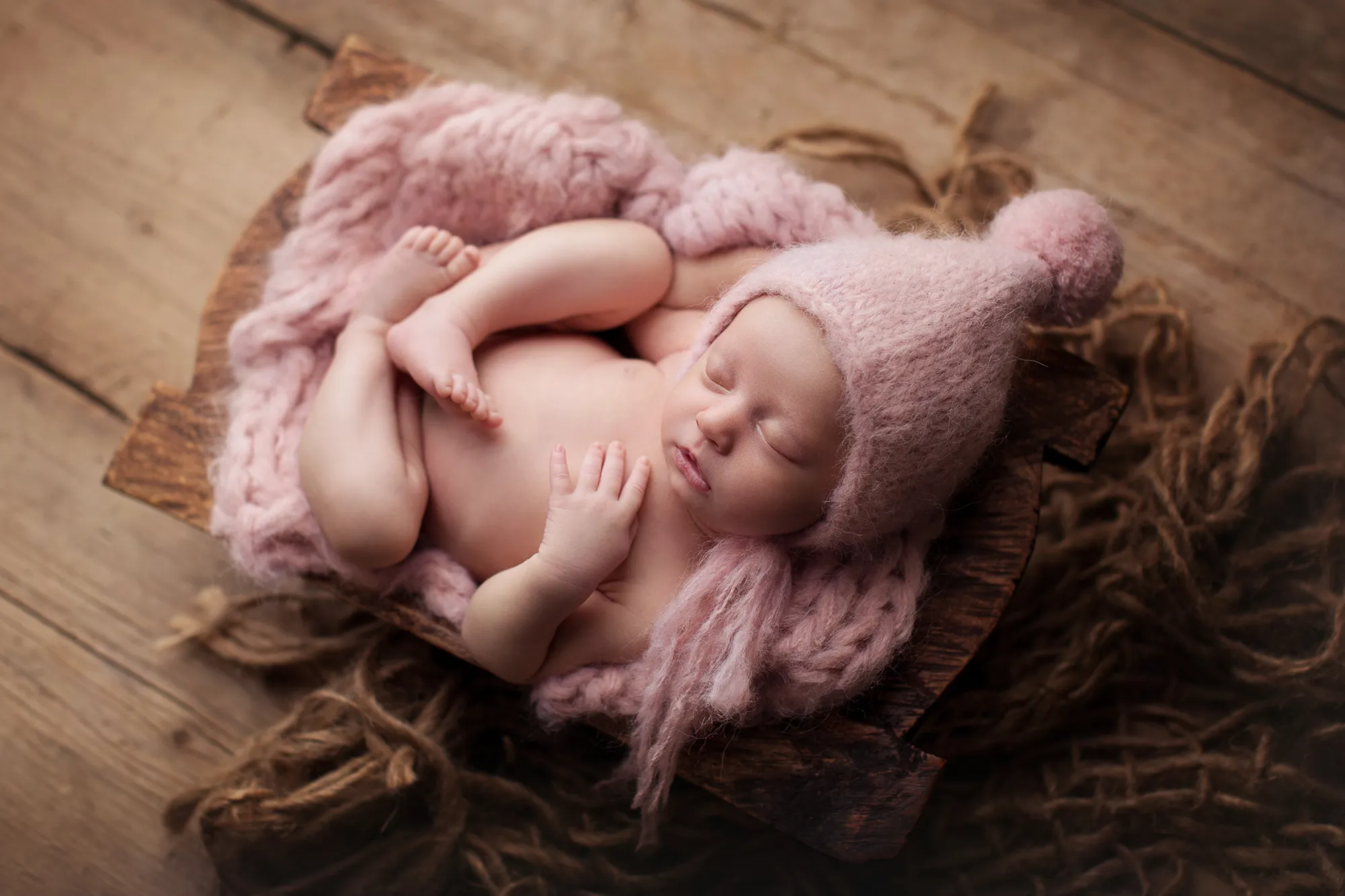 Newborn in a pink pom-pom hat asleep on a pink knit blanket in a wooden bowl
