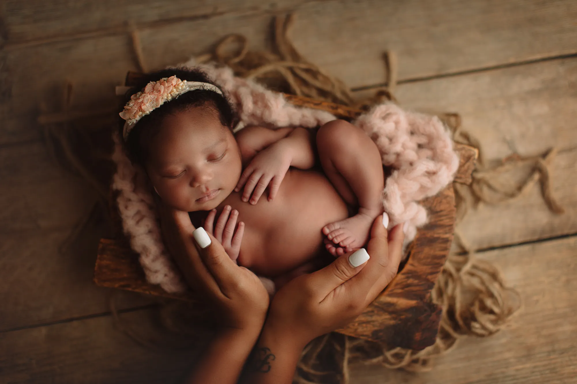 Newborn with a floral headband held in mother's hands in a rustic bowl