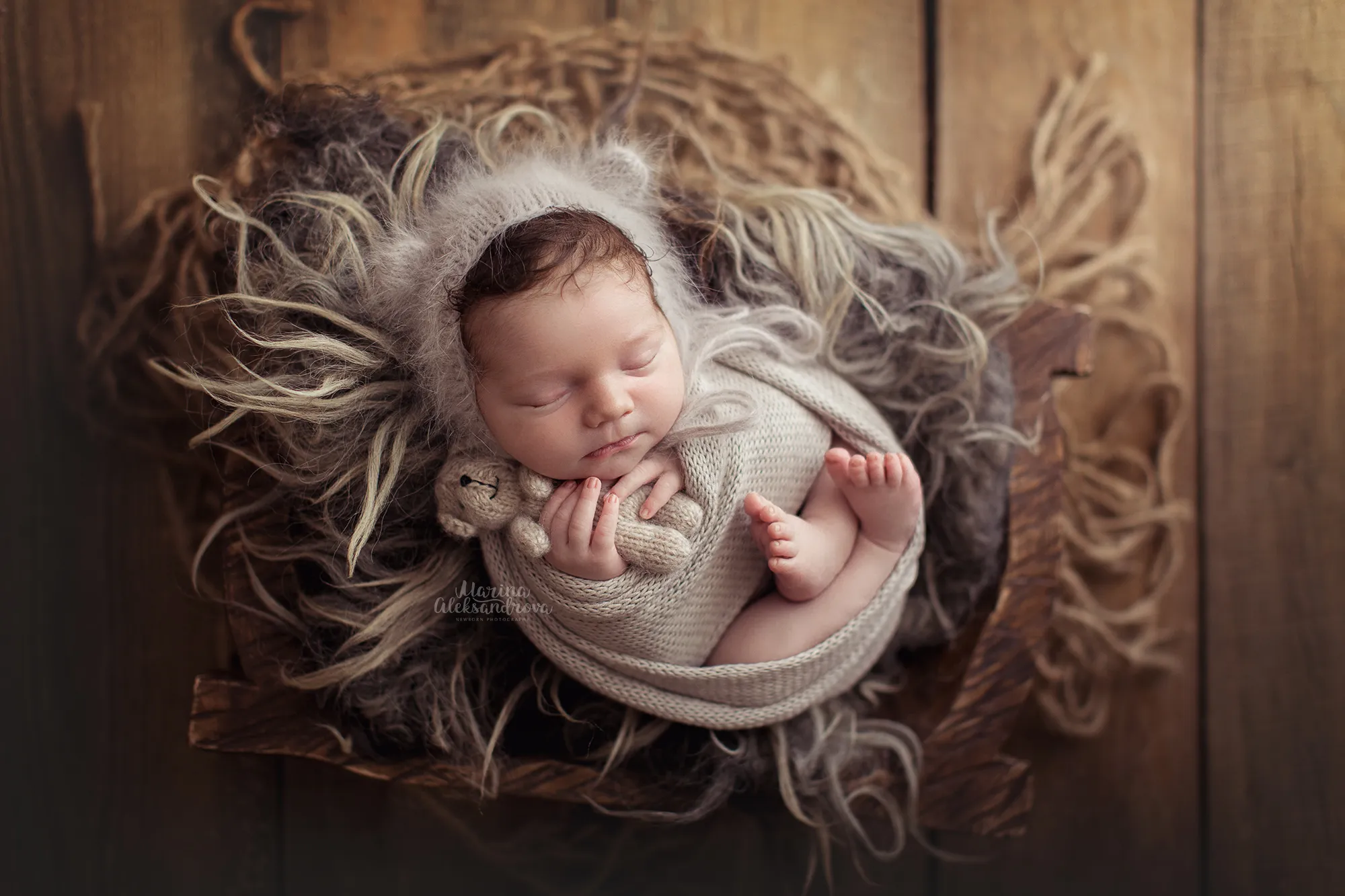 Newborn in a fluffy grey bonnet asleep in a woven nest on wooden floorboards