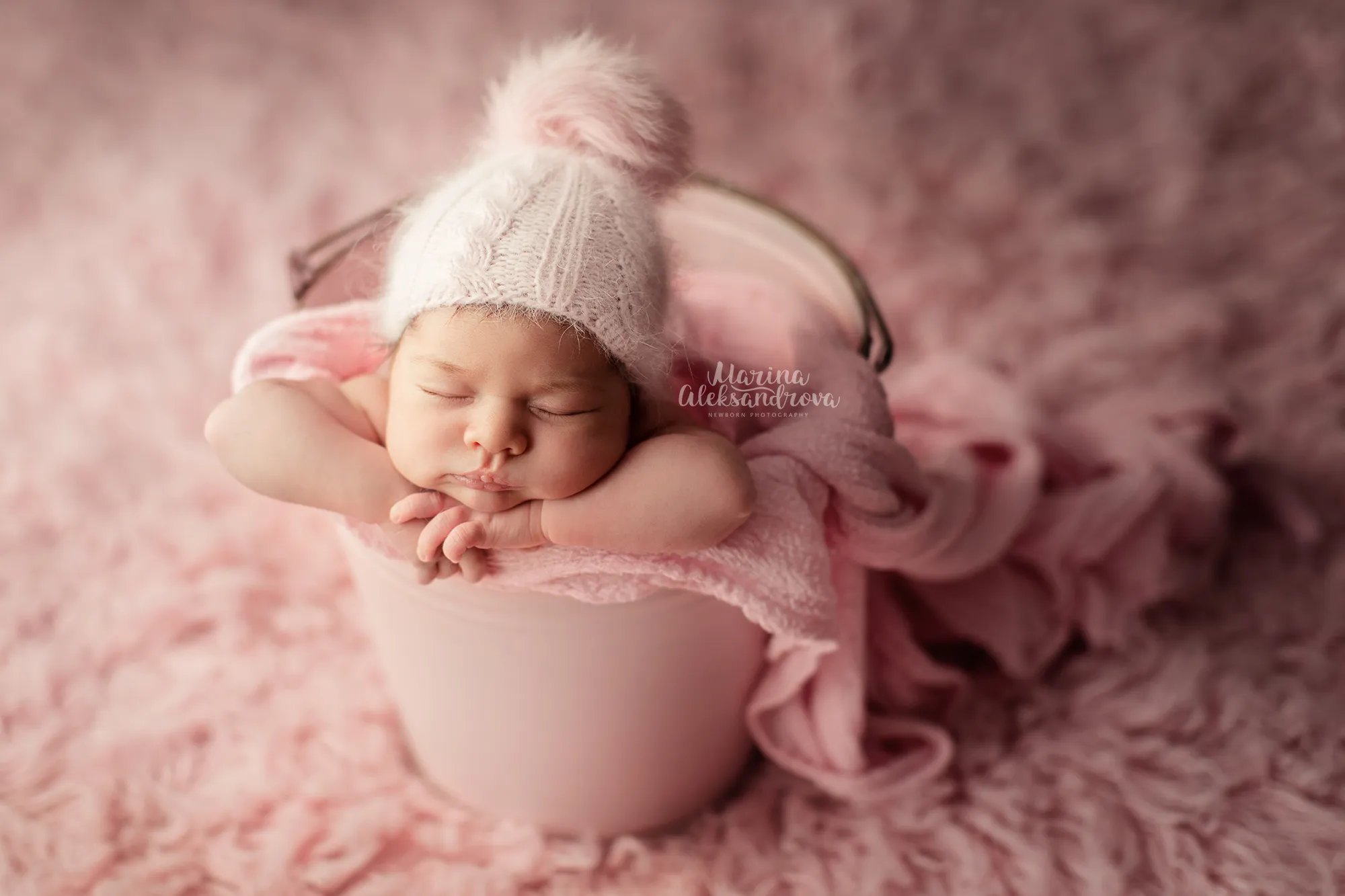 Newborn in a pink pom-pom bonnet asleep in a pink bucket on pink fur