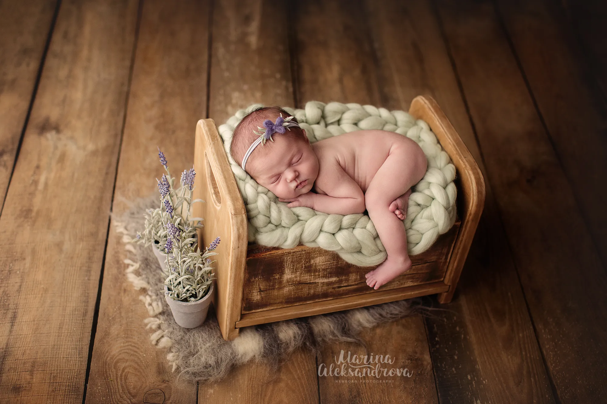 Newborn with a lavender headband asleep on a sage chunky-knit blanket in a wooden bed