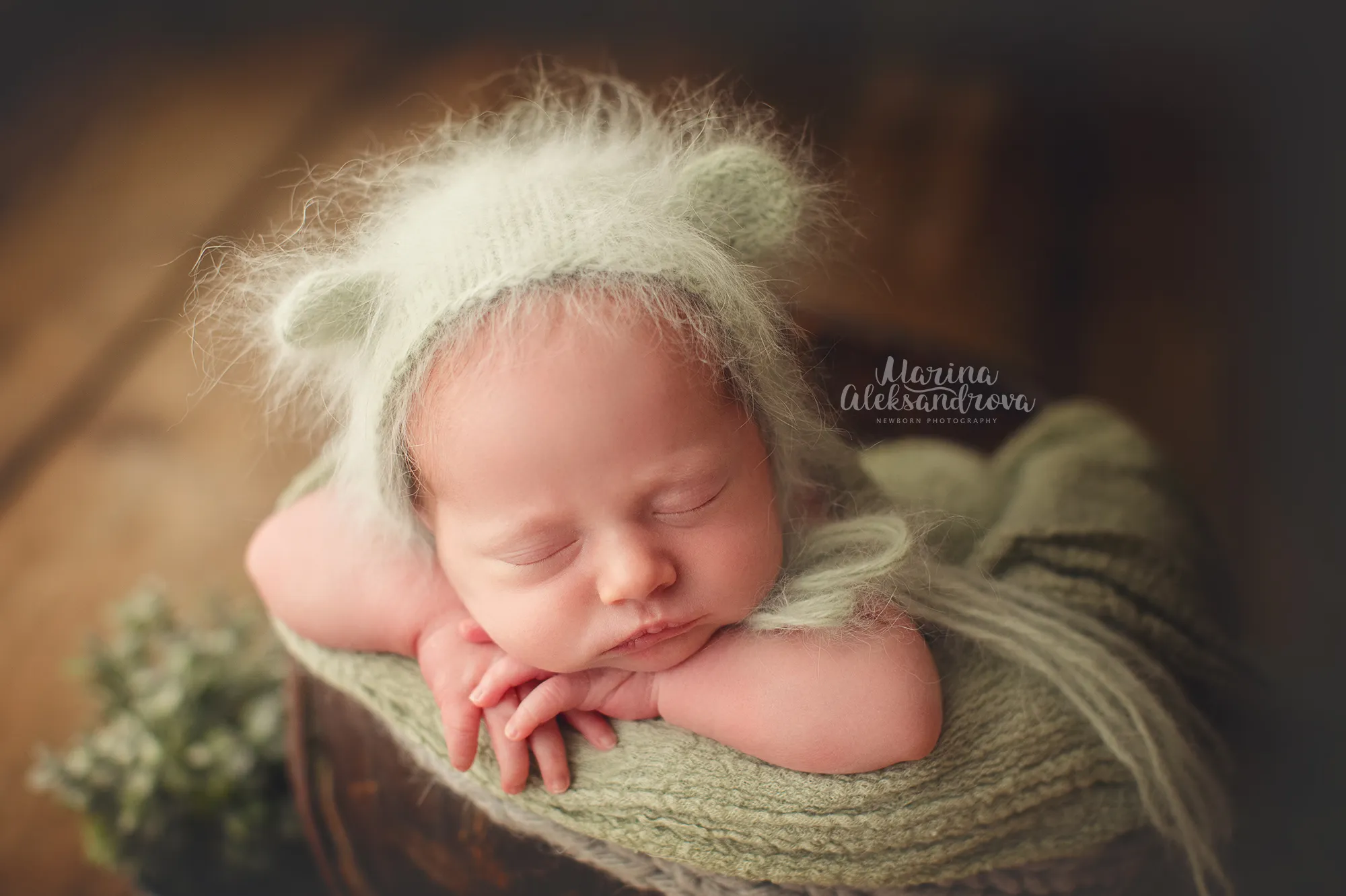 Newborn in a fluffy bear-ear bonnet resting chin on arms, sage wrap
