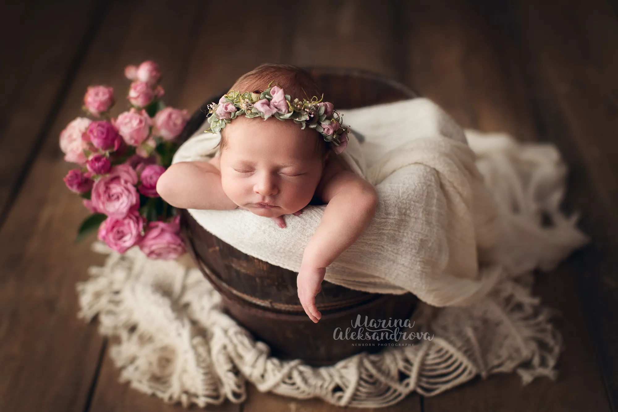 Newborn with a flower crown asleep in a bucket beside pink roses