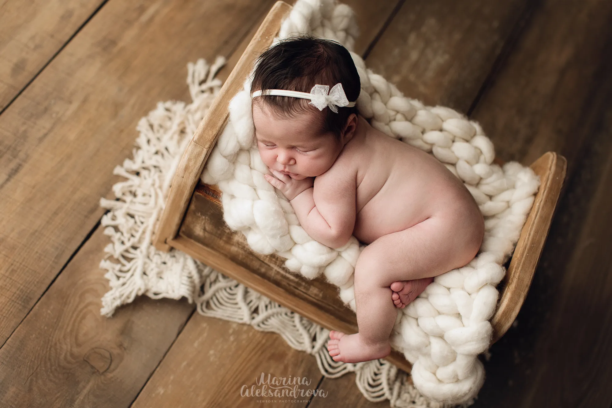 Newborn with a white bow headband asleep on a cream chunky-knit blanket in a wooden crate