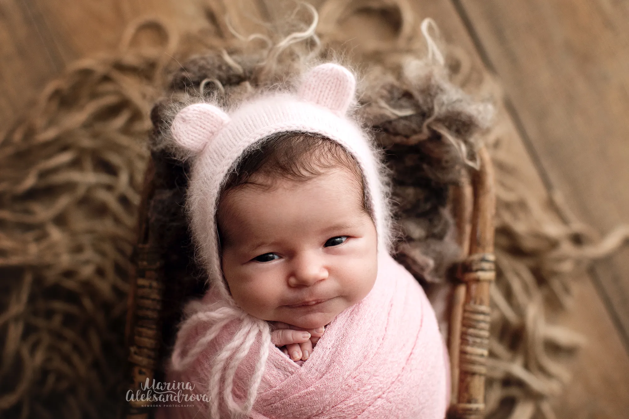 Smiling newborn in a pink bear-ear bonnet swaddled in pink in a basket