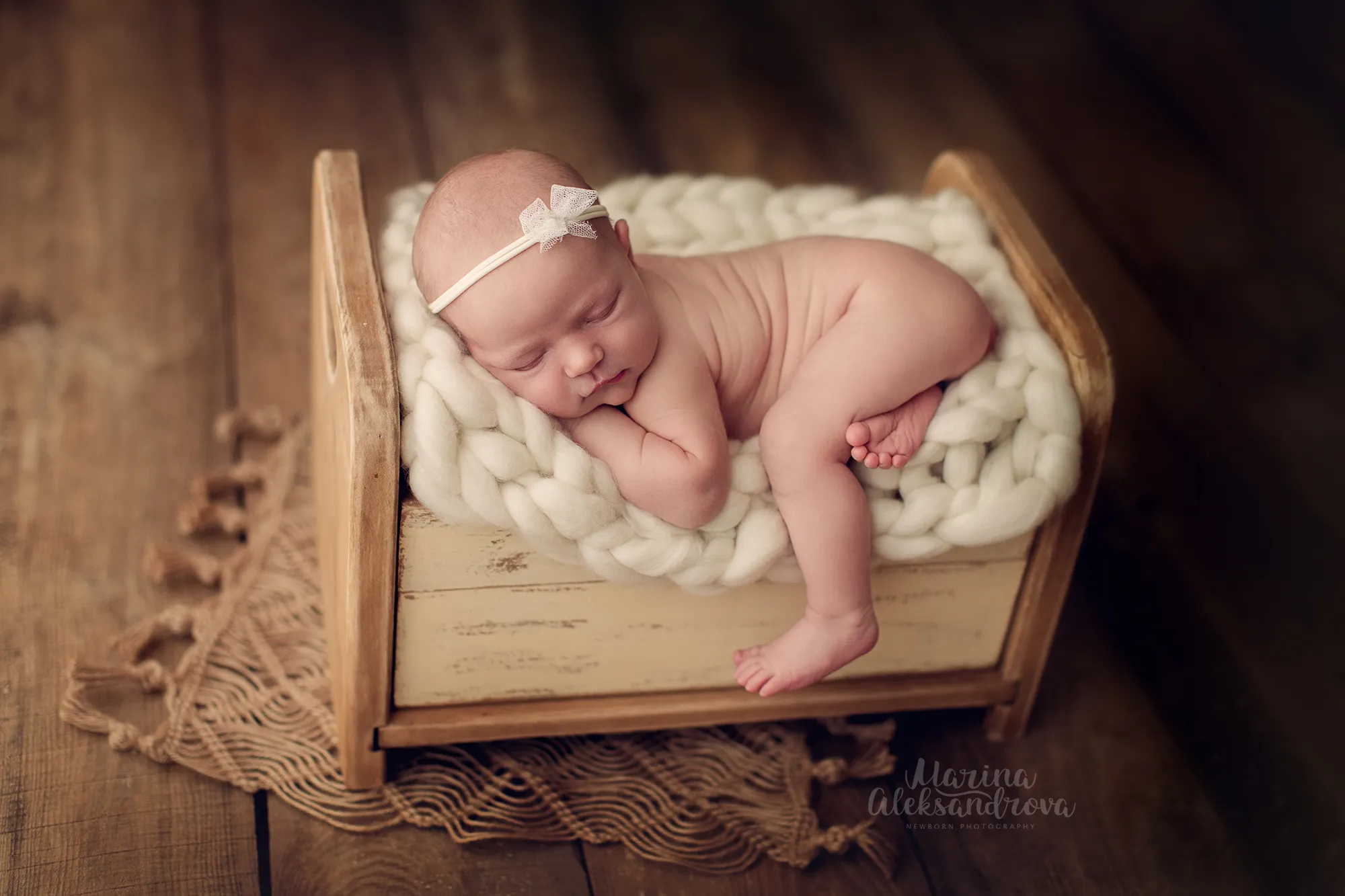 Newborn with a bow headband asleep on a cream chunky-knit blanket in a wooden bed