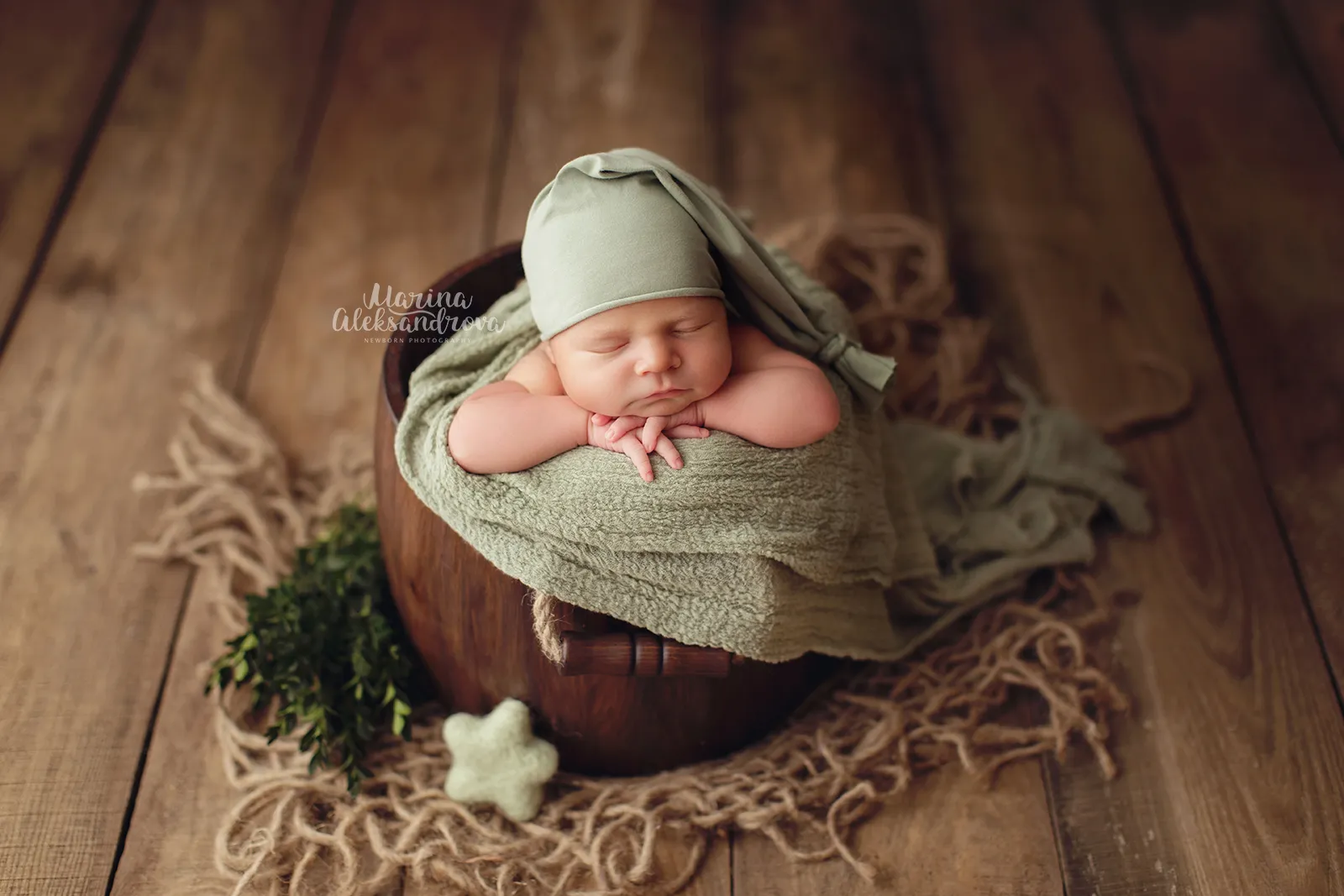 Newborn in a sage sleepy hat resting in a wooden bucket beside greenery