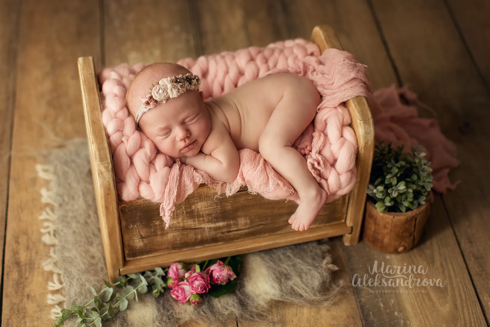 Newborn with a floral headband asleep on a pink knit blanket in a wooden bed with roses