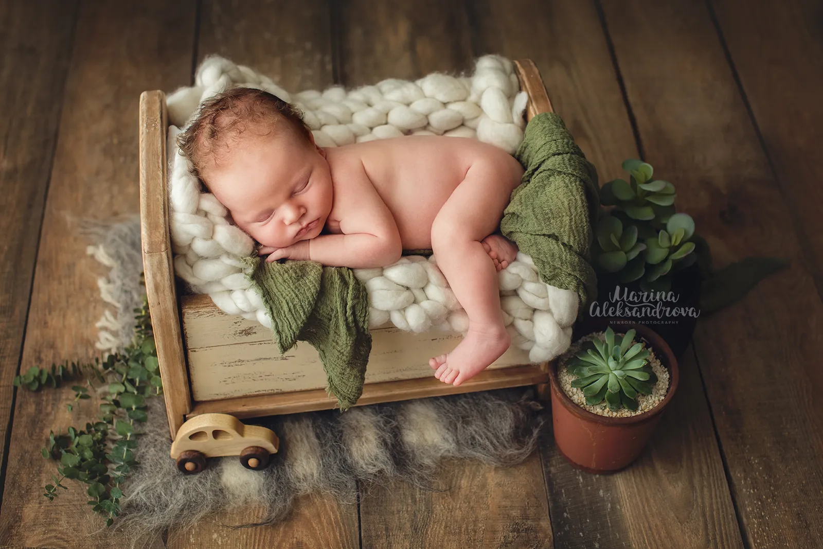 Newborn asleep on a cream chunky-knit blanket in a wooden bed with a toy car